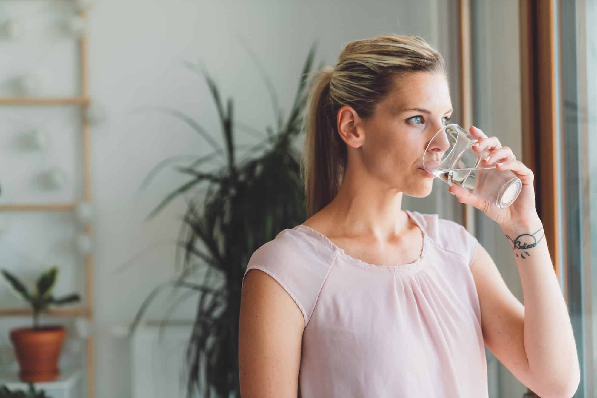 Portrait of blonde woman with a pony tail drinking a glass of water while standing by the window of